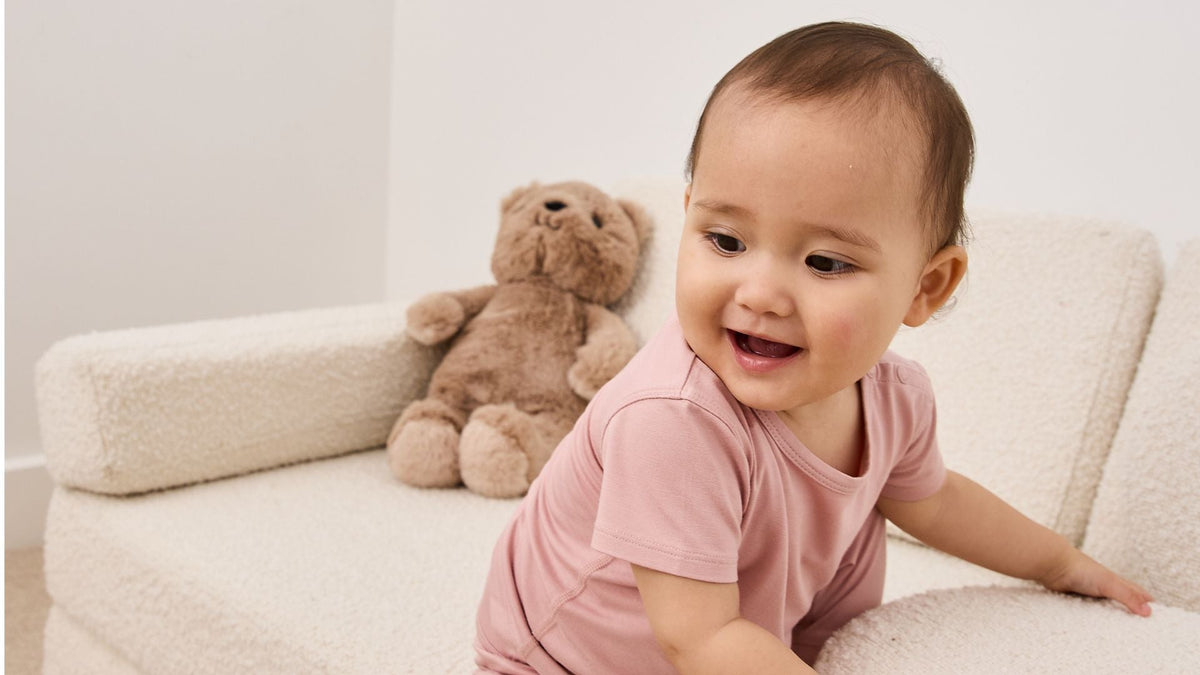 Smiling baby wearing a soft pink bamboo T-shirt sitting beside a teddy bear.